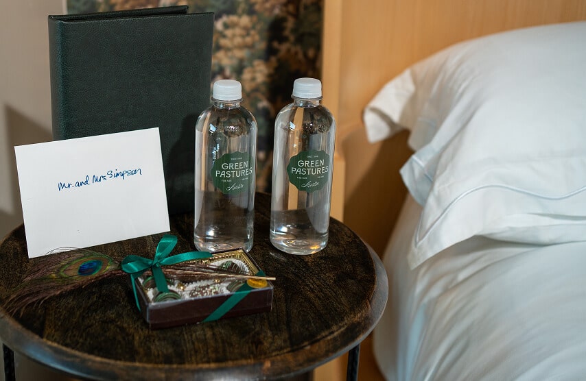 Bedside table with welcome note, bottled water, chocolates, and pillows in a luxurious hotel room.