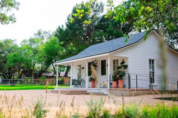White cottage with a porch surrounded by trees and greenery on a sunny day