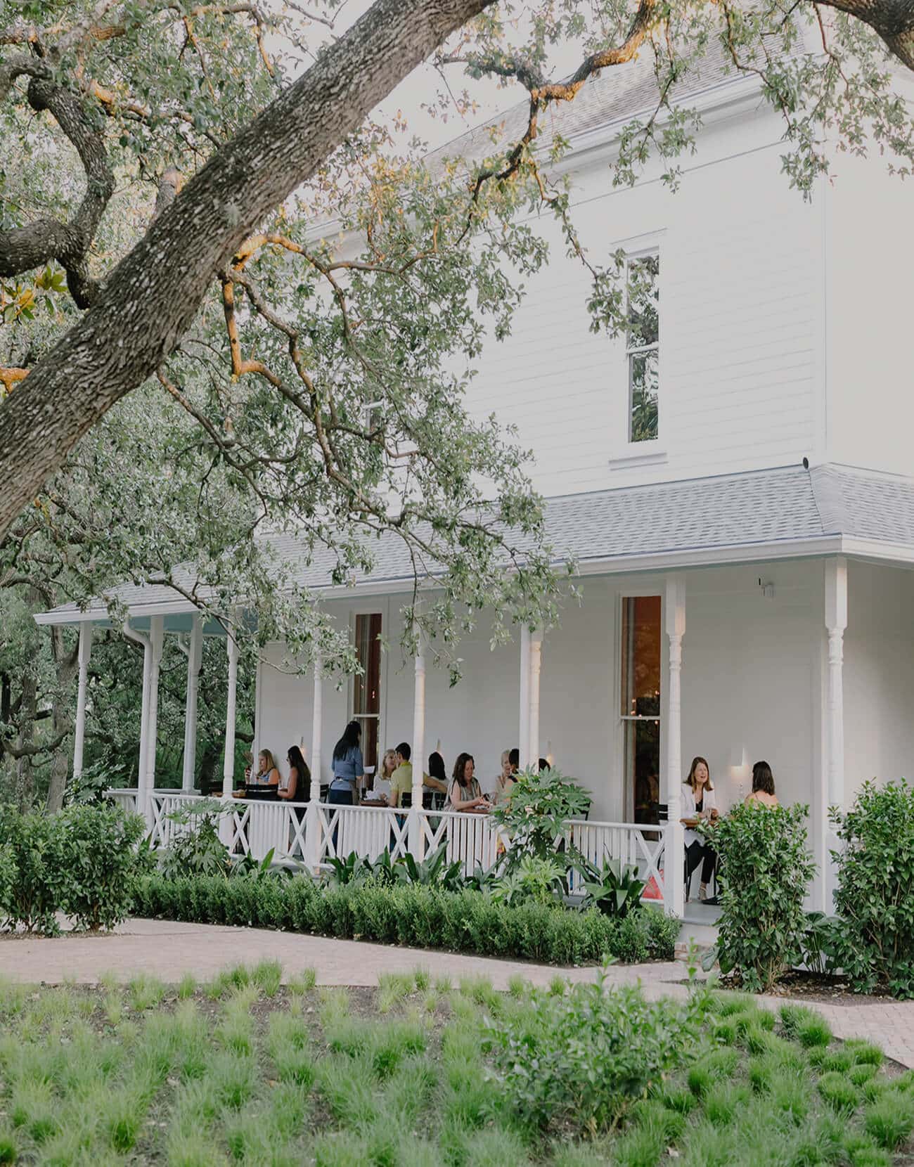 People enjoying a meal on a charming white porch surrounded by lush greenery and trees.