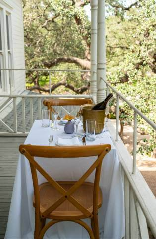 Outdoor dining on a balcony with white tablecloth and wooden chairs surrounded by trees.