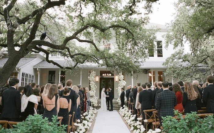 Outdoor wedding ceremony under trees with guests seated, bride and groom at altar, elegant floral decorations