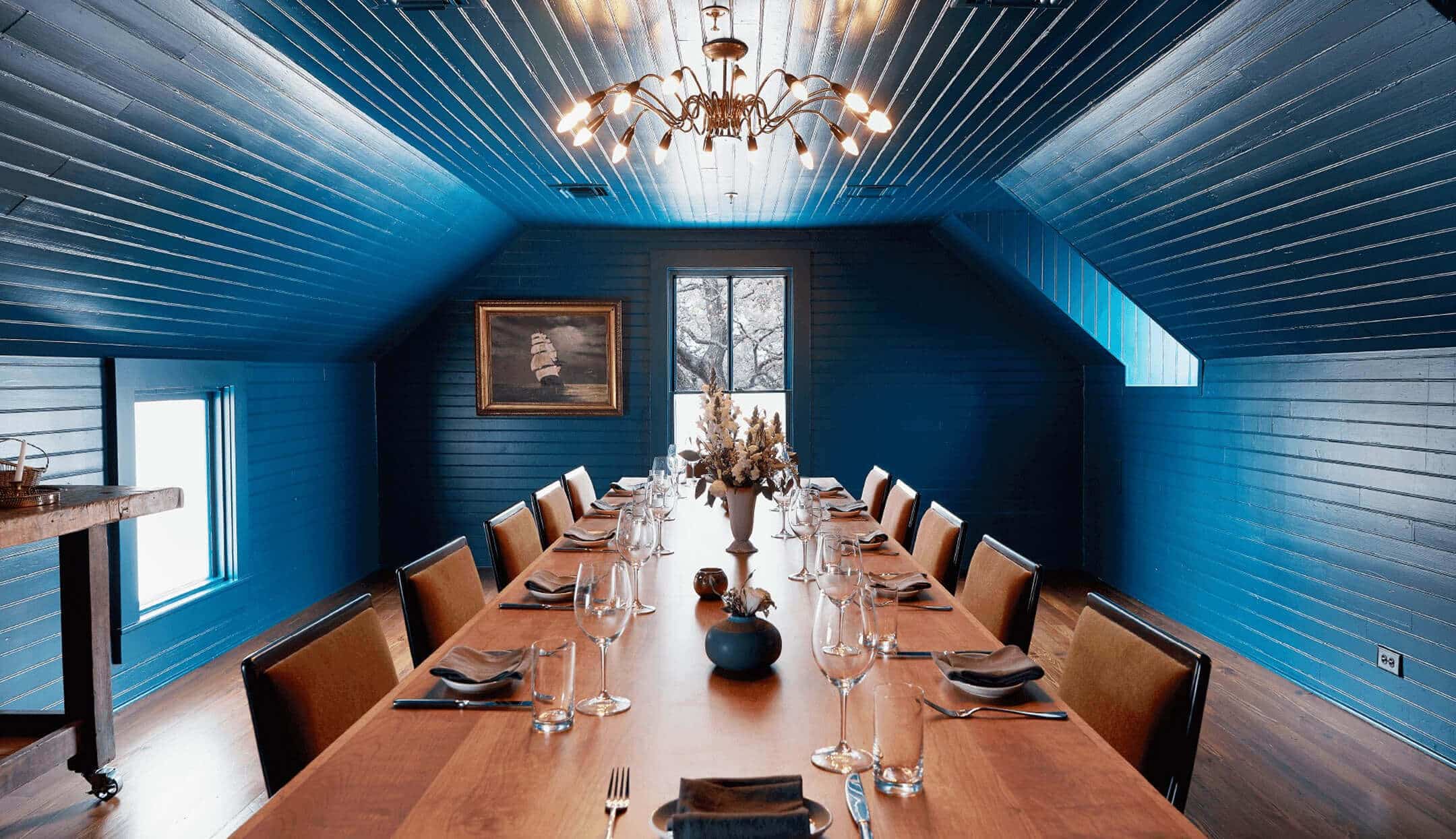 Elegant dining room with a long wooden table set for dinner under a stylish chandelier in a blue-paneled space