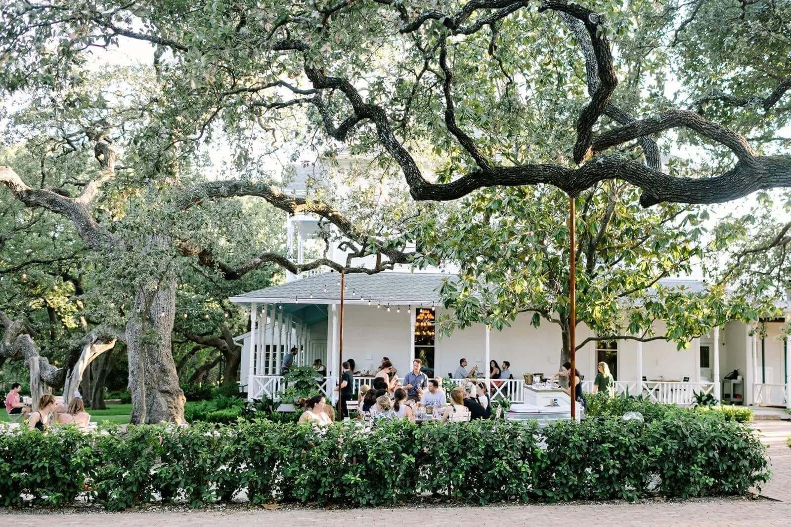 People enjoying an outdoor gathering under large trees by a charming white house
