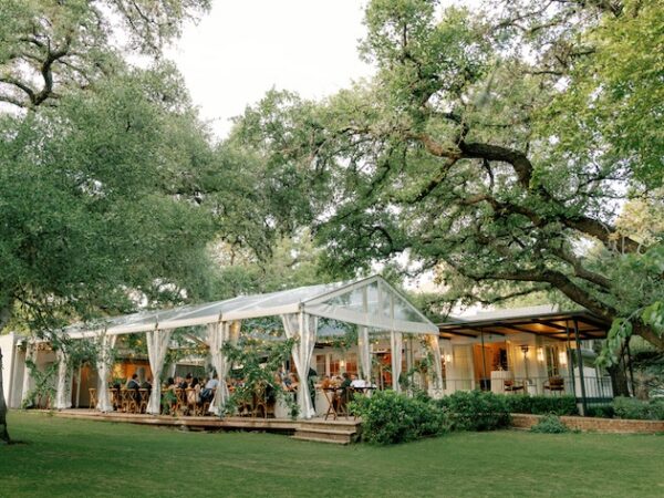Outdoor wedding reception under a clear tent surrounded by lush trees and greenery