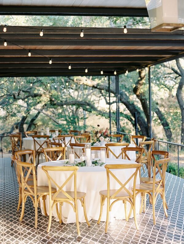 Outdoor wedding reception with round tables and wooden chairs under string lights surrounded by trees